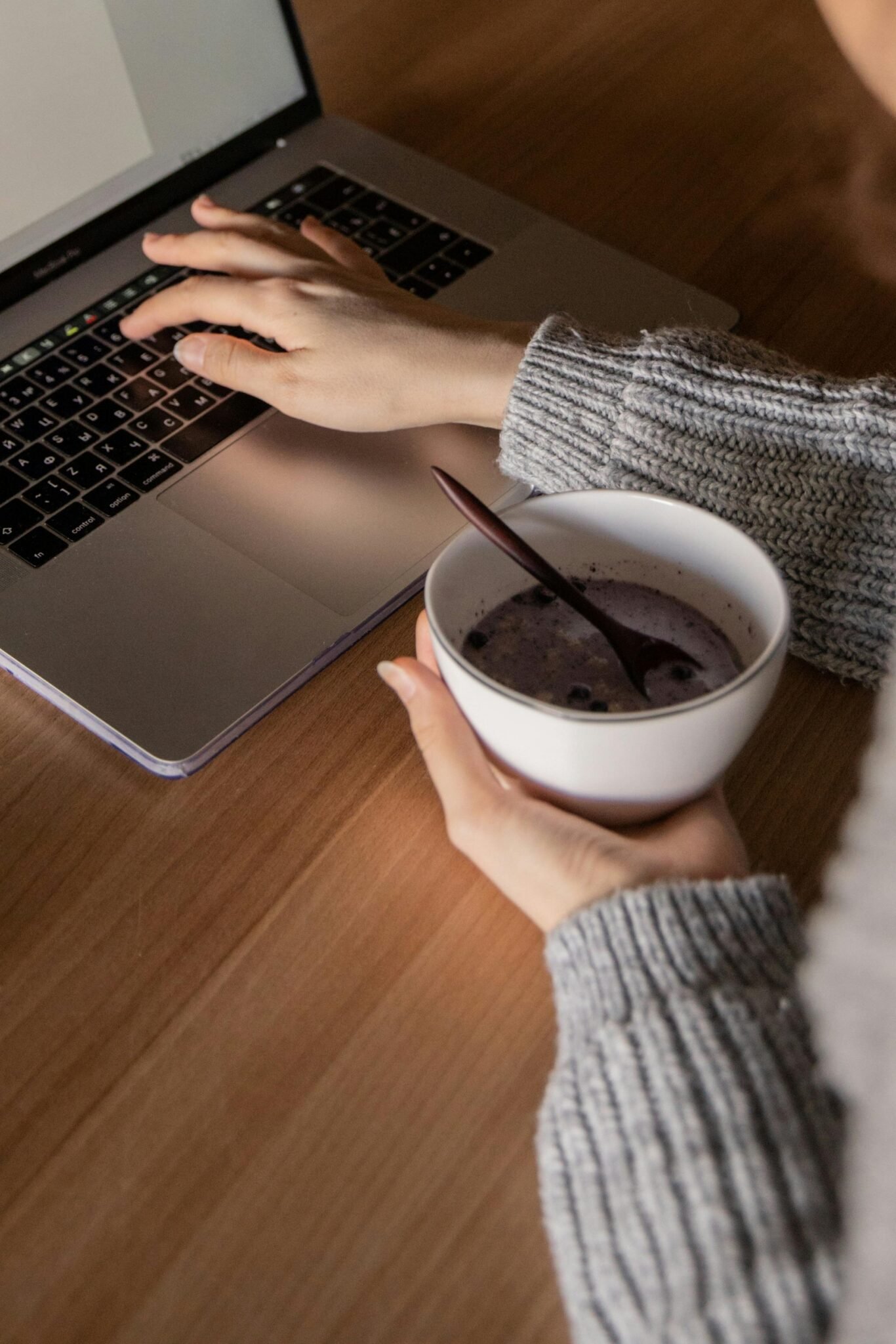 Person working on a laptop while holding a bowl of breakfast indoors.